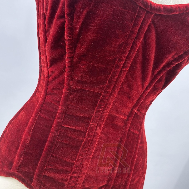 Puede incluir: Primer plano de un cors&eacute; de terciopelo rojo intenso. El cors&eacute; tiene costuras verticales y un dise&ntilde;o estructurado. La tela tiene un aspecto suave y texturizado. El cors&eacute; se muestra en un maniqu&iacute;.