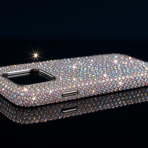 Puede incluir: Funda de teléfono adornada con pedrería iridiscente. La funda rectangular tiene bordes redondeados y un recorte para la cámara. La pedrería crea un efecto brillante. La funda se muestra sobre una superficie negra.