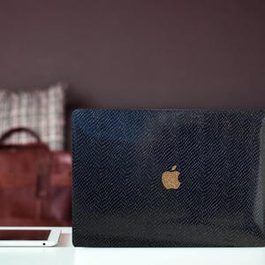 Pode incluir: Um computador portátil preto com uma superfície texturizada e um logotipo da Apple dourado. O laptop está sobre uma mesa com um tablet branco e um telefone preto.