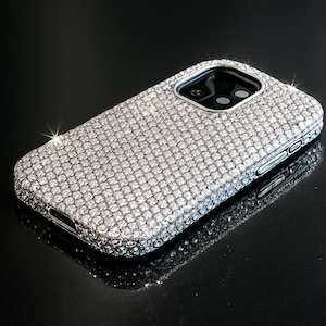 May include: A sparkling, silver phone case encrusted with numerous small, reflective crystals. The case has a camera cutout and button details, and it sits on a glossy black surface. The overall effect is luxurious and glamorous.