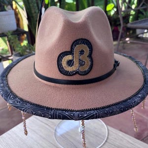 May include: A tan fedora hat featuring a black leather band and a gold embroidered logo on the front. The brim is trimmed with a black fabric featuring a white geometric pattern and dangling light brown crystal beads around the entire edge.