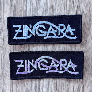 May include: Two rectangular black embroidered patches featuring the word ZINGARA in stylized lettering. The top patch has light blue embroidery, while the bottom patch features a gradient of light blue to purple thread for the text.