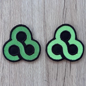 May include: Two identical embroidered patches featuring a stylized triquetra-inspired design. Each patch has a vibrant green, rounded, three-lobed shape with a black border and three circular black cutouts arranged in a central triangular pattern.
