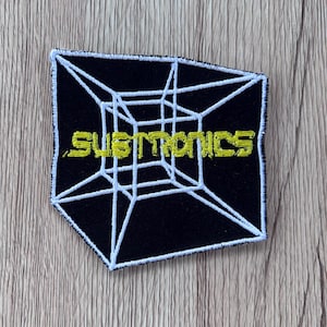 May include: A black embroidered patch featuring a white wireframe cube design and the word SUBTRONICS stitched in yellow thread across the center. The patch has a white border and a geometric shape with sharp angles and clean, bold embroidery lines.