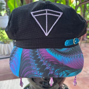 May include: A black textured newsboy cap featuring a white embroidered triangle emblem on the front, a teal velvet strap with a gold buckle, and a brim with a purple and blue fractal pattern. Three small purple crystal beads hang from the brim.