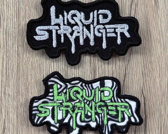 Peut inclure: Deux écussons brodés présentant le texte Liquid Stranger dans une police dégoulinante. L'écusson du haut a du texte blanc sur fond noir. L'écusson du bas a du texte vert citron sur un fond à motif marbré noir et blanc.