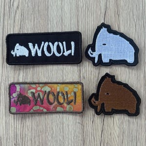May include: Four embroidered patches featuring woolly mammoth designs. Two are rectangular with the text "WOOLI" and a small mammoth icon, one in black and white, the other in a multicolored tie-dye pattern. Two are shaped like mammoths, one light blue and one brown.