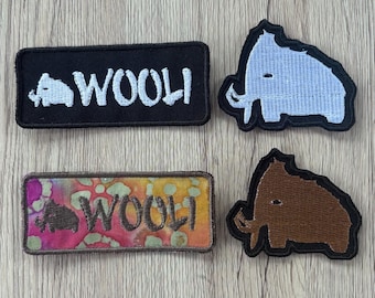 Peut inclure: Quatre écussons brodés représentant des mammouths laineux. Deux sont rectangulaires avec le texte "WOOLI" et une petite icône de mammouth, l'un en noir et blanc, l'autre avec un motif tie-dye multicolore. Deux ont la forme de mammouths, l'un bleu clair et l'autre marron.