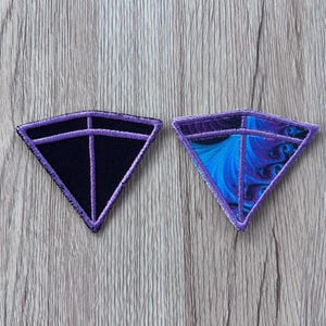 May include: Two triangular embroidered patches with purple stitched borders and internal lines. One patch features a solid black center, while the other displays a vibrant blue and purple fractal pattern. Both patches have a distinct geometric shape.