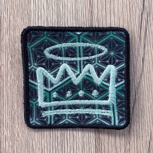May include: A square embroidered patch featuring a light blue crown and halo design centered over a dark green and black geometric hexagonal pattern. The patch has a thick black stitched border and a flat, fabric-like texture throughout the design.