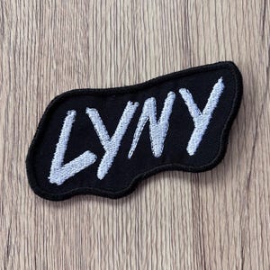 May include: A black embroidered patch with an irregular, wavy-edged shape featuring the text LYNY stitched in white thread. The lettering has a hand-drawn, slightly slanted appearance, and the entire patch is outlined with a thick black border.
