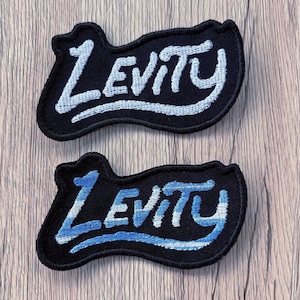 May include: Two black embroidered patches with irregular, wavy edges. Each patch features the word Levity stitched in a stylized, cursive font. The top patch uses white thread, while the bottom patch uses a gradient of light blue and white thread.