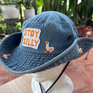 May include: A blue denim bucket hat featuring an embroidered white patch with the orange text STAY SILLY. The hat is decorated with small embroidered white goose figures with orange beaks and feet, and includes a dark blue adjustable chin strap.