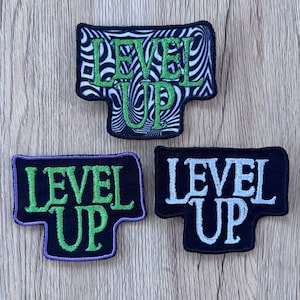 May include: Three embroidered patches featuring the text LEVEL UP. The top patch has a black and white wavy pattern with green text. The bottom left patch is black with green text and a purple border. The bottom right patch is black with white text.