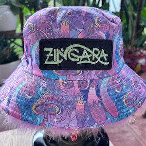 May include: A bucket hat featuring a vibrant purple and blue mushroom pattern with a black rectangular patch on the front displaying the text ZINGARA in light green embroidery. The brim is trimmed with light pink faux fur and beads.
