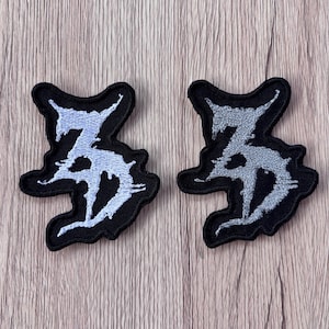 May include: Two embroidered patches featuring a stylized, jagged, black-bordered design with a light gray, distressed-looking symbol in the center. The patches have an irregular, organic shape with sharp, pointed edges and a dense, textured thread finish.
