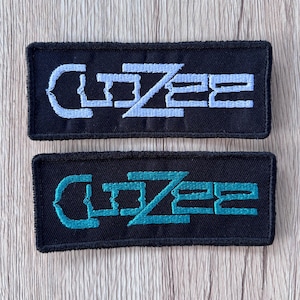 May include: Two rectangular black embroidered patches featuring the text CLOZEE in a stylized font. The top patch has light blue embroidery, while the bottom patch features teal embroidery. Both patches have a black stitched border edge.