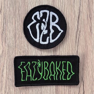 May include: Two embroidered patches on a wood grain surface. The top patch is circular and black with white stylized letters forming the initials EZB. The bottom patch is rectangular and black with green embroidered text reading EAZYBAKED.