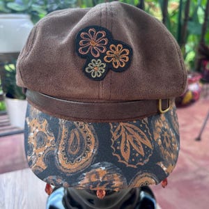 May include: A brown suede newsboy cap featuring a dark brown leather band with a gold-tone buckle. The front brim displays a black fabric with a tan and brown paisley pattern. An embroidered patch of three flowers sits on the crown, and small amber-colored beads hang from the edge of the brim.