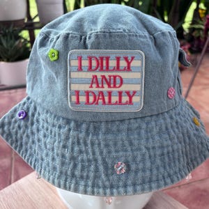 May include: A light blue denim bucket hat featuring a rectangular patch with horizontal stripes and the embroidered red text I DILLY AND I DALLY. The hat is decorated with various small colorful buttons and clear beads attached to the brim.