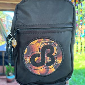 May include: A black nylon sling bag featuring a circular patch with a swirling orange, brown, and yellow pattern and a black embroidered logo in the center. A small, clear, hexagonal crystal-shaped charm hangs from the front zipper pull.