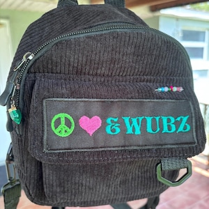 May include: A black corduroy mini backpack featuring a rectangular patch on the front pocket. The patch displays an embroidered green peace sign, a pink heart, and the teal text "& WUBZ". A small row of colorful beads is attached above the patch.