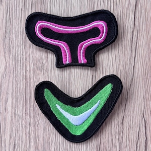 May include: Two embroidered patches with black fabric bases. The top patch is a black horizontal shape with a bright pink outline. The bottom patch is a black V-shape featuring a green chevron design with a white accent line in the center.