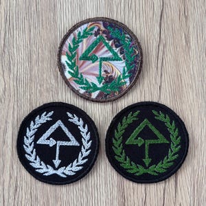 May include: Three circular embroidered patches featuring a central arrow symbol pointing upward and downward, enclosed by a laurel wreath. One patch has a multicolored marbled background, while the other two have solid black backgrounds with silver or green stitching.