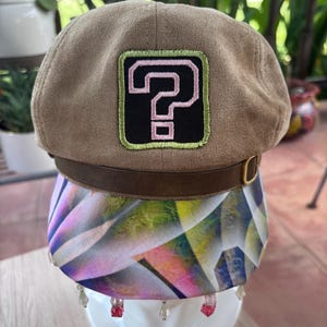 May include: A tan suede newsboy cap featuring a central patch with a black background, a light green border, and a pink question mark. The cap includes a dark brown leather-like band with a gold buckle and a multicolored geometric patterned brim.