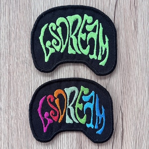 May include: Two black, crescent-shaped embroidered patches. The top patch features the word "LSDREAM" stitched in bright lime green thread. The bottom patch displays the same word "LSDREAM" with each letter in a different color: pink, orange, light green, and blue.