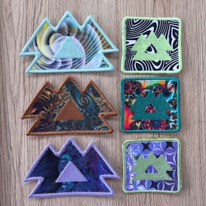 May include: Six embroidered patches arranged in two columns. The left column features three mountain-shaped patches with swirling patterns in blue, brown, and purple. The right column contains three square patches with geometric and fractal designs.