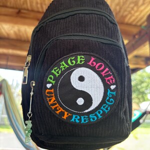 May include: A black corduroy sling backpack featuring a circular embroidered patch with a central yin-yang symbol. The patch text reads PEACE LOVE UNITY RESPECT in multicolored thread. A small flower pin is at the top and a green crystal charm hangs from the zipper.
