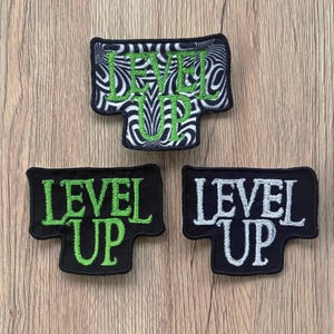 May include: Three embroidered patches with the words "LEVEL UP". One patch has a black and white swirled background with green lettering. The other two patches are black with green and white lettering.