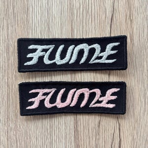 May include: Two rectangular black patches with the word "FUME" embroidered in a cursive font. One patch has white embroidery, and the other has pink embroidery. The patches are displayed on a light brown wooden surface.
