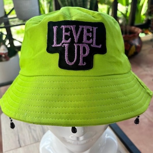 Puede incluir: Un sombrero de pescador verde neón con las palabras "LEVEL UP" en letras moradas sobre un parche negro. El sombrero tiene un ala ancha y adornos negros en forma de lágrima. El sombrero se exhibe en una cabeza de maniquí blanca.