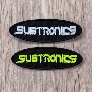 May include: Two oval-shaped embroidered patches featuring the text SUBTRONICS in a stylized font. One patch has white lettering on a black background, while the other patch features bright neon green lettering on a black background.