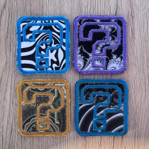 May include: Four square embroidered patches, each featuring a central question mark outline. The top-left patch has a blue border and black-and-white swirl pattern. The top-right has a purple border and black-and-white fractal design. The bottom-left has a gold border and paisley pattern. The bottom-right has a blue border and black-and-white wave pattern.