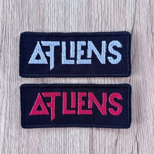 May include: Two rectangular embroidered patches with black fabric backgrounds. The top patch features the text ATLIENS in light blue thread, while the bottom patch displays the same text in bright pink thread. Both patches have black borders.