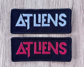 Peut inclure: Deux écussons rectangulaires brodés sur fond de tissu noir. L'écusson du haut présente le texte ATLIENS en fil bleu clair, tandis que celui du bas affiche le même texte en fil rose vif. Les deux écussons ont des bordures noires.