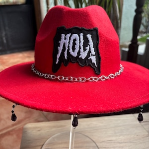 Puede incluir: Un sombrero fedora rojo con una cadena plateada alrededor de la corona y detalles de cuentas negras. El sombrero presenta un parche negro con la palabra "HOLA" en letras blancas. El sombrero se exhibe sobre un soporte transparente.