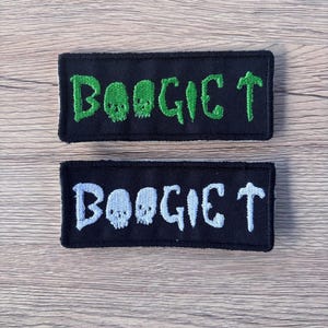 May include: Two rectangular black patches with the word "BOOGIE T" embroidered in green and white. The letter "O" in "BOOGIE" is replaced with a skull design. The patches are on a light brown wooden surface.