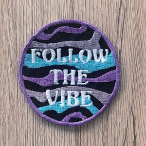 May include: Round embroidered patch with the text "FOLLOW THE VIBE" in white. The patch has a black, purple, and teal wave pattern and a purple border. The patch is on a wooden surface.