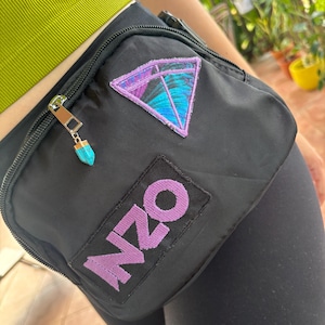 Puede incluir: Riñonera negra con un parche triangular morado y turquesa y la palabra "NZO" en morado. El tirador de la cremallera tiene un pequeño dije turquesa. La riñonera se lleva alrededor de la cintura.