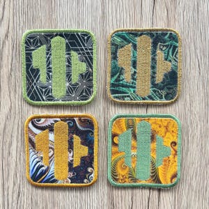 May include: Four square embroidered patches with a central design. Each patch features a different color scheme: green on black, gold on green, gold on blue, and green on yellow. The patches have a geometric and abstract design.