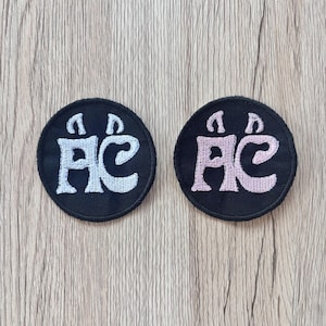 May include: Two black, circular patches with embroidered white and pink Japanese characters. The patches are on a light brown wooden surface. The characters are stylized and appear to be a symbol or logo.