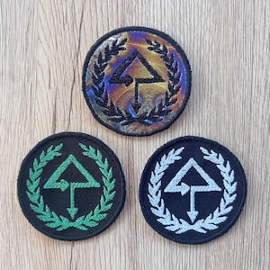 Puede incluir: Tres parches redondos bordados con un diseño geométrico. El parche superior tiene un fondo iridiscente multicolor con un borde negro y una corona de laurel negra. Los otros dos parches son negros con un diseño y borde verde y blanco.