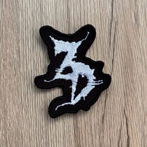 May include: An embroidered patch featuring a black background with a white, jagged, stylized letter design. The patch has a thick black border and an irregular, organic shape that follows the contours of the white embroidered lettering.