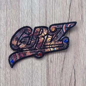 May include: Embroidered patch with the word "GRRL" in a stylized font. The patch features a paisley-like pattern in shades of brown, orange, and blue, with a black border. It is embellished with small, colorful gems.