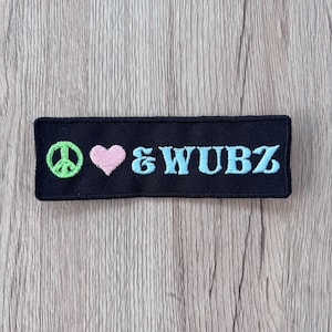 May include: A rectangular black embroidered patch featuring a lime green peace sign, a light pink heart, and the light blue text &WUBZ. The patch has a black stitched border and is designed for application onto fabric surfaces as a decorative element.
