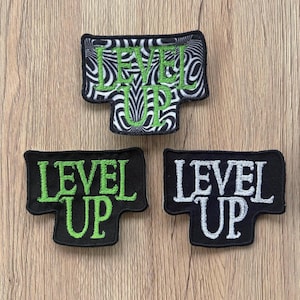Puede incluir: Tres parches bordados con el texto "LEVEL UP". Un parche tiene un fondo en espiral en blanco y negro con texto verde. Los otros dos parches son negros con texto verde y blanco.